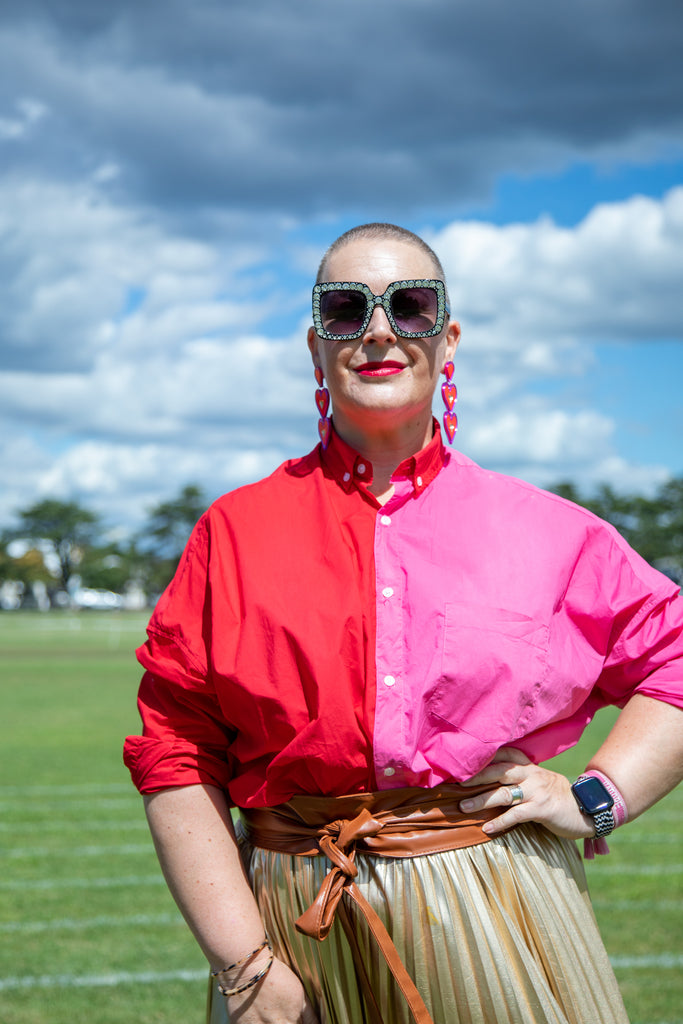 Women standing with hand on hip, with a closely cropped shaved head, wearing a red and pink shirt with Elton John like sunglasses and a gold skirt. 