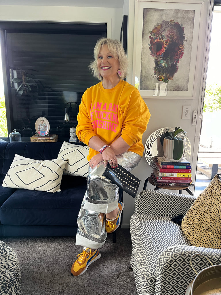 Woman sits on chair in silver pants with bright sweatshirt, smiling at camera