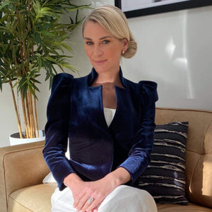 A blonde woman with a chic side knot hairstyle,  sits with legs crossed  on a couch, looking directly at camera.  She is looking down the lens. 
